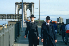 Zwei junge Männer mit Hüten, Schlips, Kragen und schwarzen Mänteln kommen zu Fuß von der Brooklyn Bridge